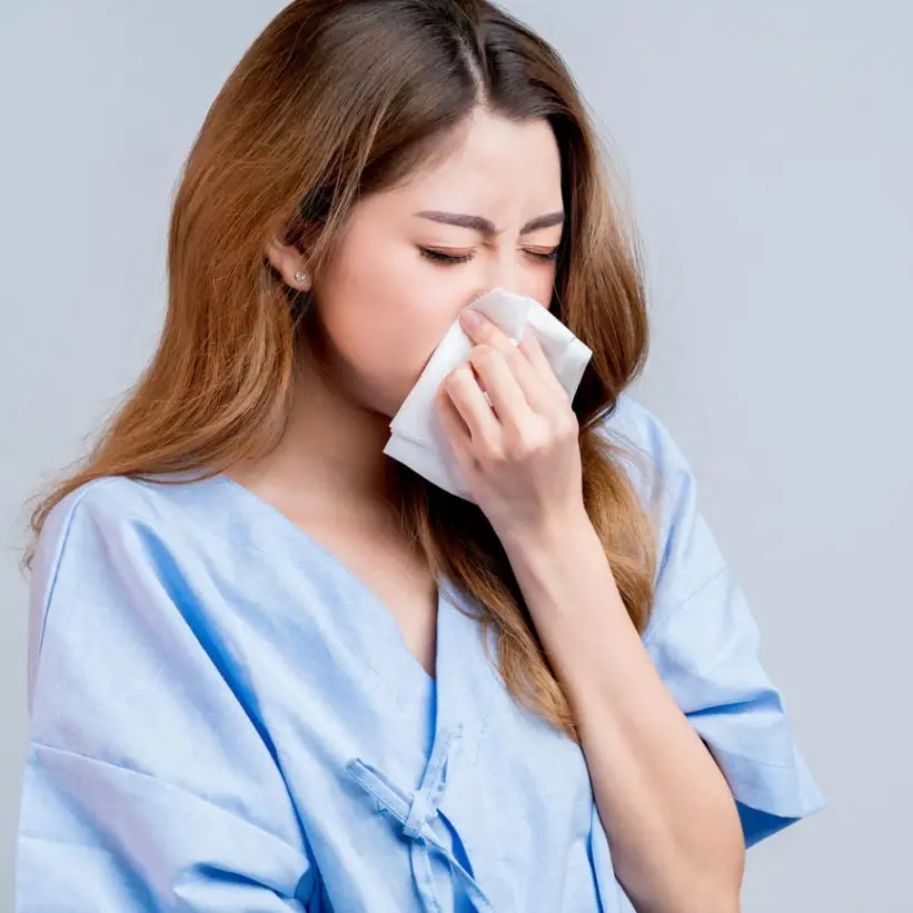 Young woman in a blue blouse sneezing into a tissue against a light gray background.