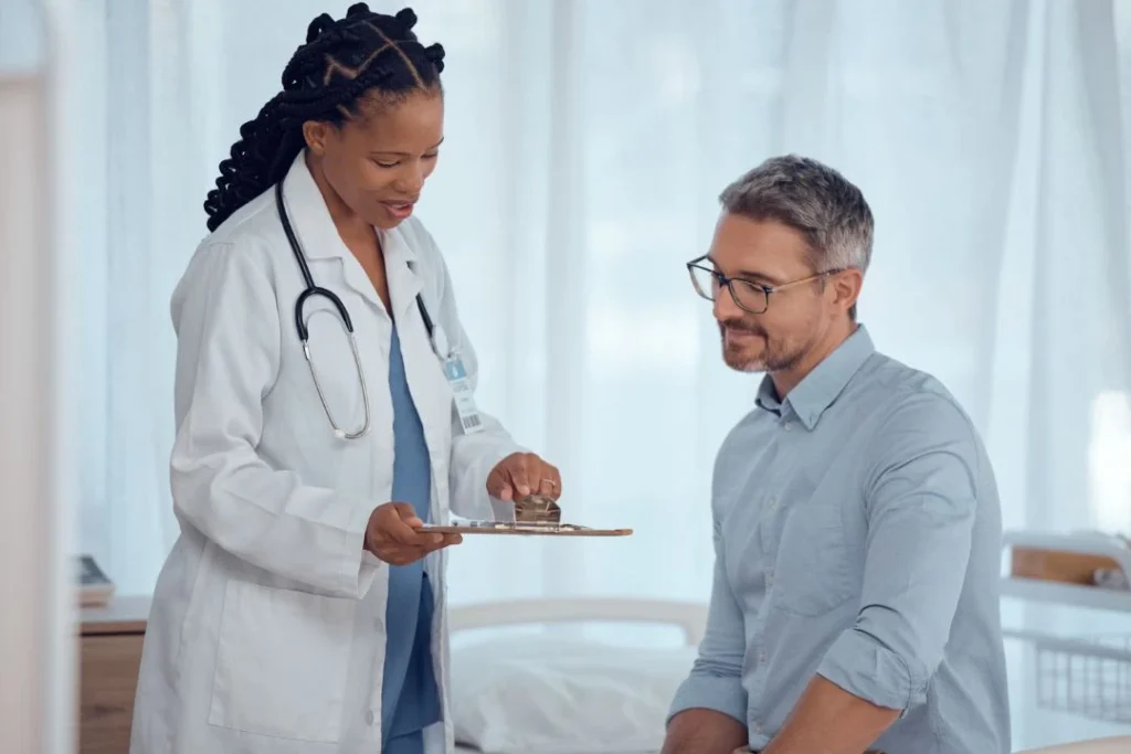 Doctor holding a clipboard & talking to a seated male patient.