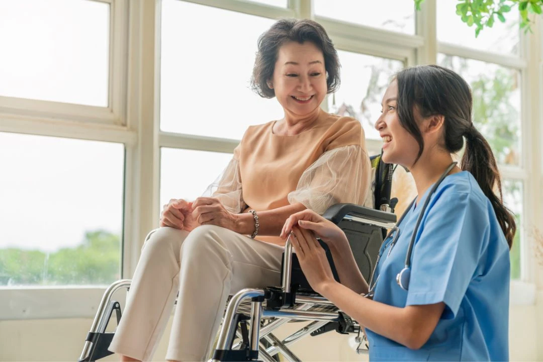 Nurse smiling & talking to a woman in a wheelchair near a bright window.